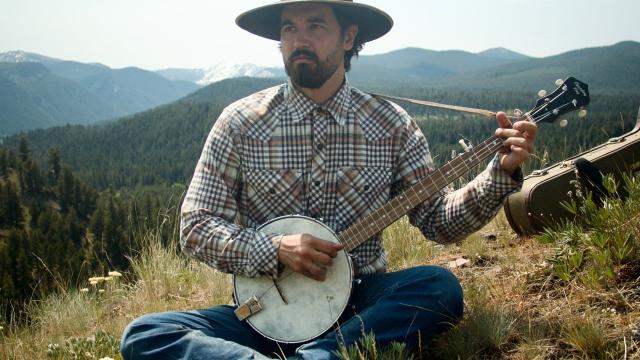 A man is wearing a hat and seating in a mountain setting and playing music 