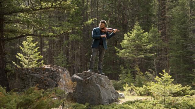 Musician Barry Shiffman standing on a rock in nature playing violin. 