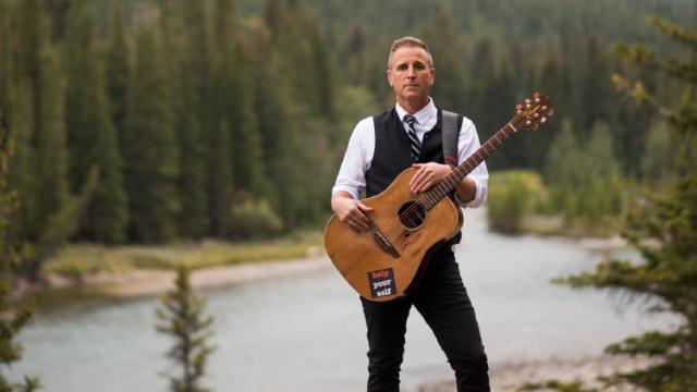 Sean McCann poses with guitar in front of a forest with a river. 