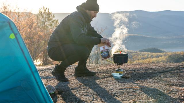 A man crouches by his tent holding a bag of Happy Yak dehydrated food and boiling water on a camp stove. His camping loc