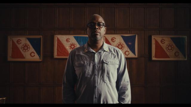 Dark-skinned man with glasses wearing a light blue shirt is standing in front of a blue white and red flag  hanging off a wood wall.