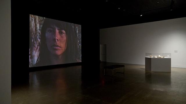 A dark gallery has a diffuse spotlight on an artistic stand, a screen in the background shows the face of an indigenous 