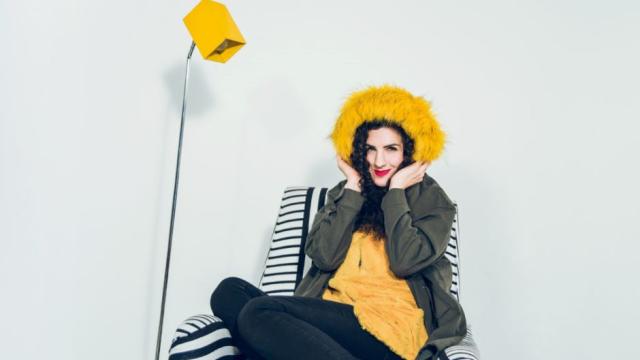 Laila Biali is wearing a bright yellow shirt and matching fur hat, while sitting on a black and white striped armchair, 
