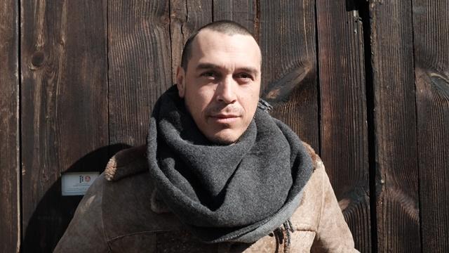 Artist Cannupa Hanska Luger stands in front a wooden wall, he is wearing a scarf and winter clothes.