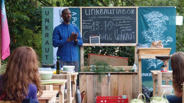 A man stands at the front of an informal outdoor classroom lecturing on Unkraut