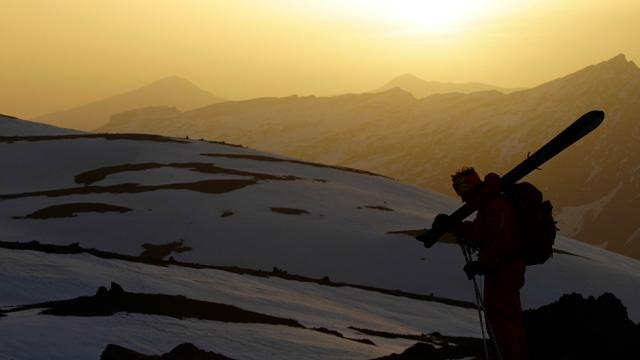 Silhouetted skier with skis resting on shoulder stands against a backdrop of snow-covered mountains as the setting sun c