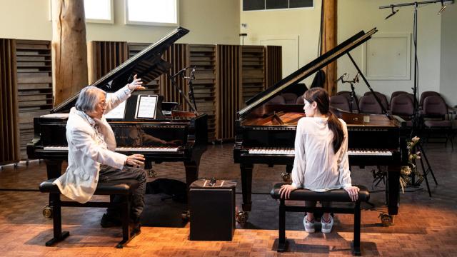 Two people are seated at a pair of grand pianos