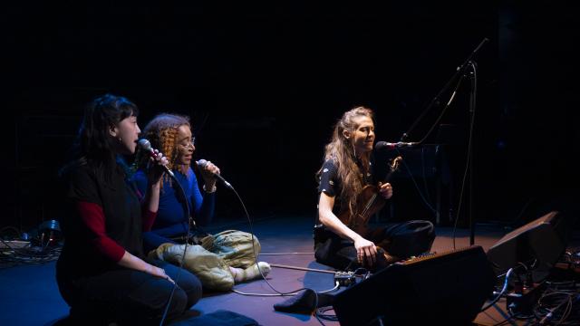 A trio of musicians are seated on the floor of a darkened stage