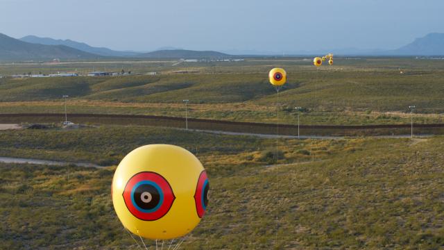 Yellow helium balloons with an eye symbol are spread out across an open landscape