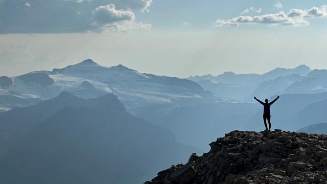 A younf woman stands on a rock outcrop of a peak, her hand extend upwards in a celebratory manner. Glaciers and icefields of the Canadian Rockies can be seen behind her.