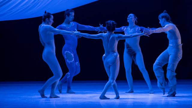Five dancers are posed in a circle with their arms outstretched and touching, on a stage that is illuminated with blue l