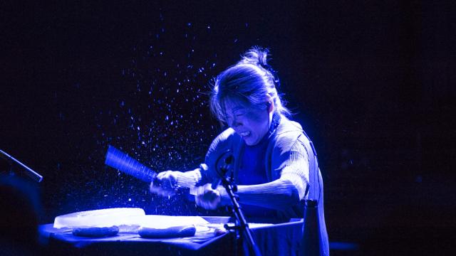 A woman lit with an overhead blue light beats a unique drum set.