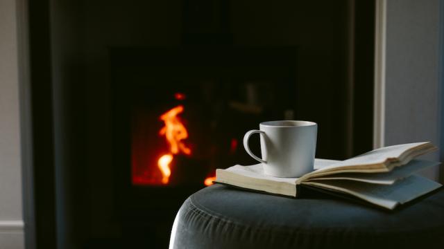 Coffee cup sitting on a book, resting upon a stool in front of a fireplace. 