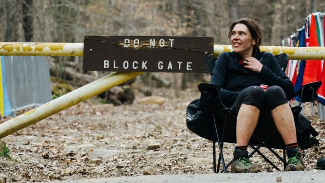 Woman wearing sport clothes and running shoes is seating in a camping chair in front of a trail head with an old wood sign indicating ''Do not block Gate''