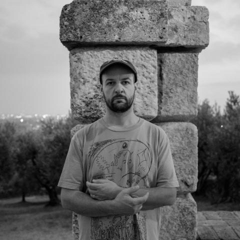 Man wearing t-shirt and hat standing in front of a stone pillar with arms folded in front of him