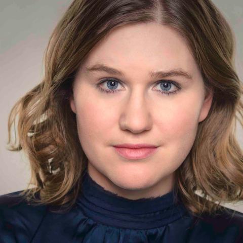 Woman with blonde shoulder length hair wearing blue satin blouse staring directly at camera and slightly smiling