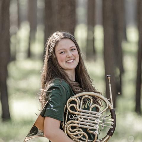 Woman standing in forest holding french horn and smiling at camera