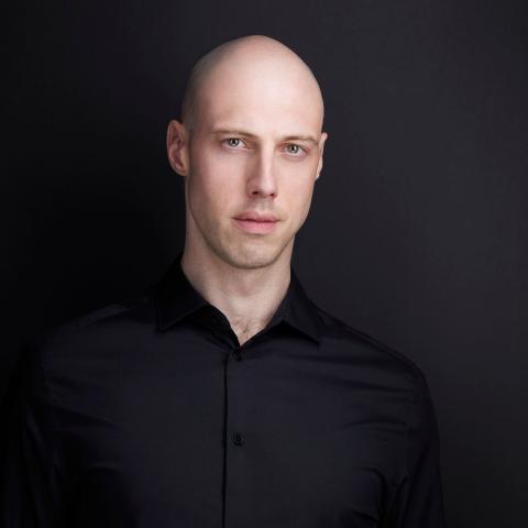 Man in black shirt standing in front of black wall staring intensely at camera