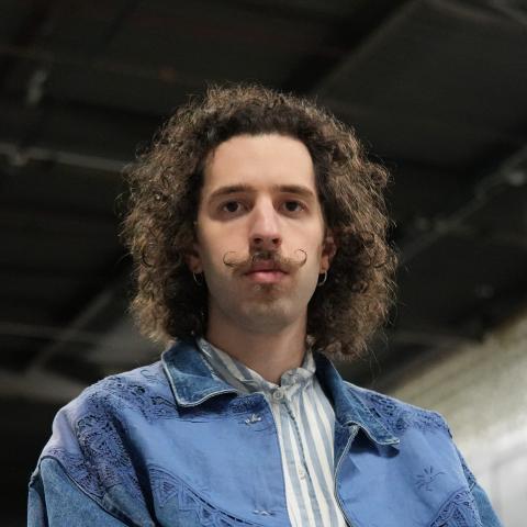 Man with shoulder length curly hair looking at camera wearing a jean jacket and a distinct moustache