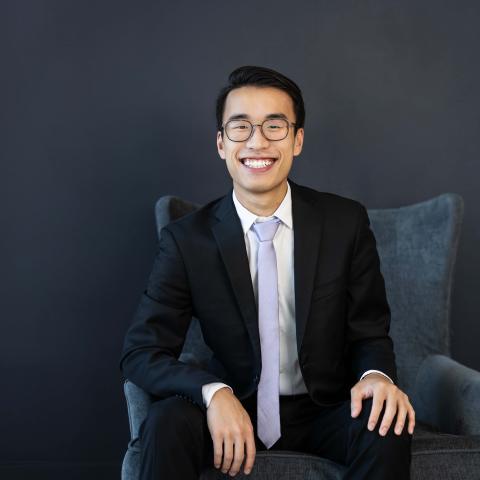 man wearing glasses and a dark suit with a light tie sitting in a comfy chair smiling a large smile at the camera