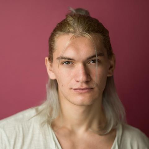 Man with shoulder length blond hair staring at camera with slight smile in front of pink backdrop