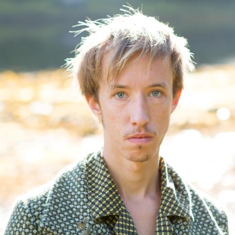 A man standing in a field in a green patterned shirt and jacket making direct eye contact with the camera