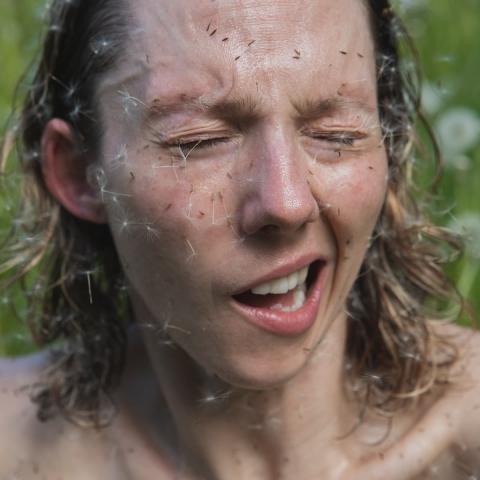 Erin has her hair down and dandelion fluffs stuck to their face. They squint. 