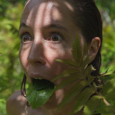 Genevieve has slicked back hair and is outdoors. There are green shrubs behind them. They have their mouth open, tongue 