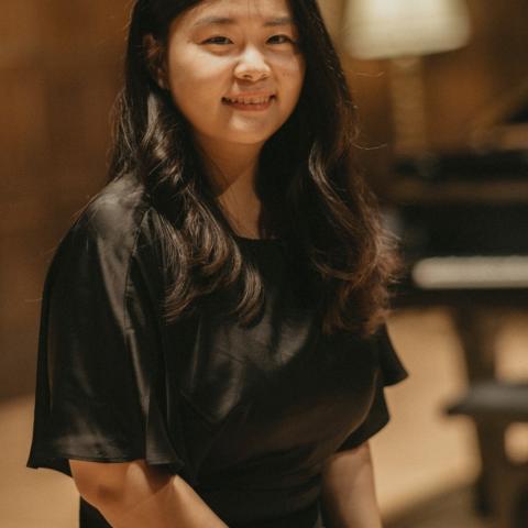 Woman sitting with hands on her lap in a formal black dress and long flowing hair over her shoulders. Piano in backgroun