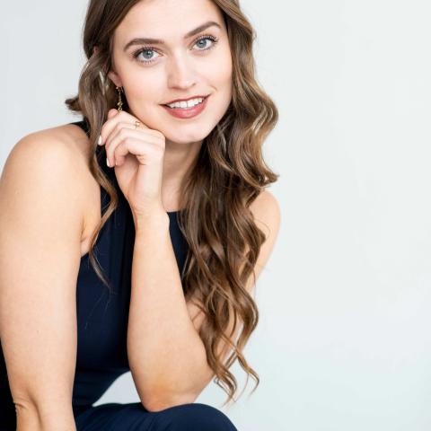 Woman with long wavy hair smiling at the camera in a black formal dress while leaning over with her elbow on her knee