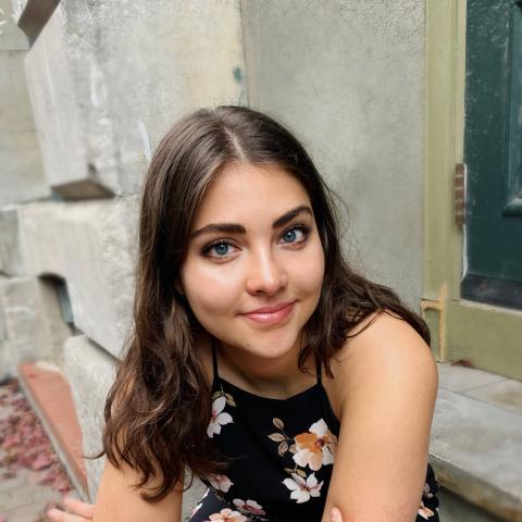 Woman with long flowing brown hair smiling at the camera sitting outside on a doorstep in a floral dress
