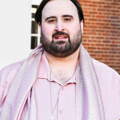 Bearded man with short dark hair looking straight at the camera wearing a pink shirt and a shiny pink scarf