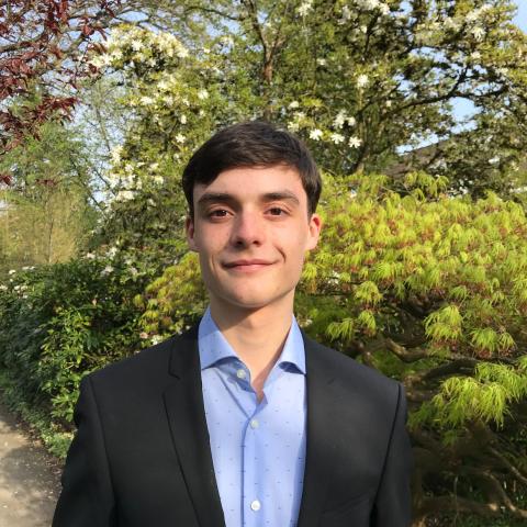 Man in dark jacket and blue dress shirt smiling directly at camera outside with trees in the background