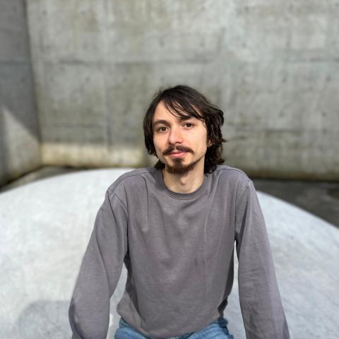 Casually dressed man in a grey long sleeved shirt and jeans sitting outside in front of a concrete wall and smiling at t