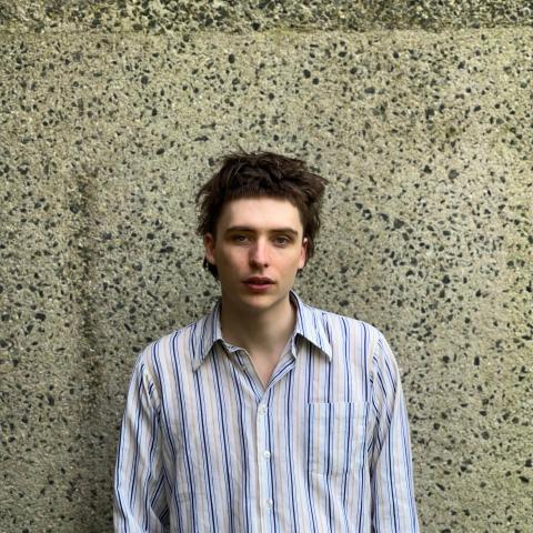 Short haired man in purple striped shirt standing against a wall and looking into the camera
