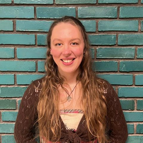 Woman with long blond hair smiling at the camera leaning against a green brick wall