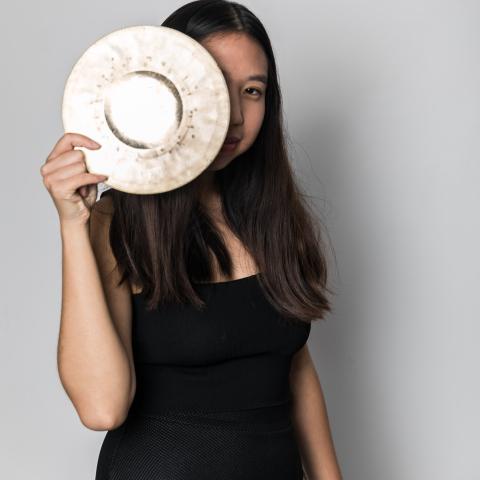 Woman smiling behind a cymbal she's holding in front of her face and peeking out from
