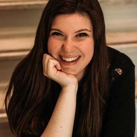 Woman smiling at the camera with her hand on her chin sitting on some stairs which rise in the background