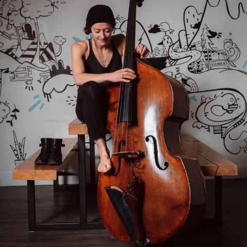 Jill sits in front of a white wall with black drawing art on the wall. She plays an upright base and wears a black toque