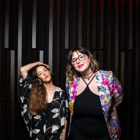 Marie-Clo on the left and éemi on the right pose in front of a striped background. Marie-Clo wears a dark outfit with wh