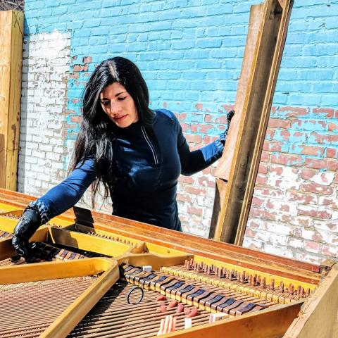 Nicole stands in front of a blue and white rustic brick wall playing the inside of a piano. 