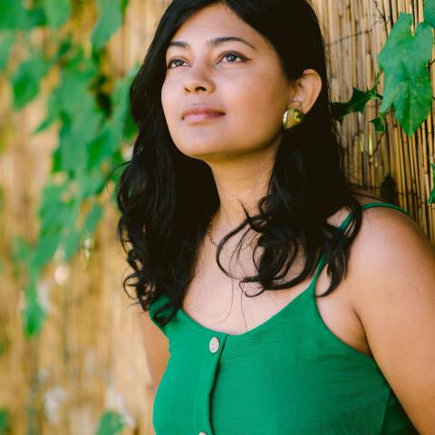 Priyanka is in a green dress against a naturesque and fenced background. She stares off into the left of the frame. 
