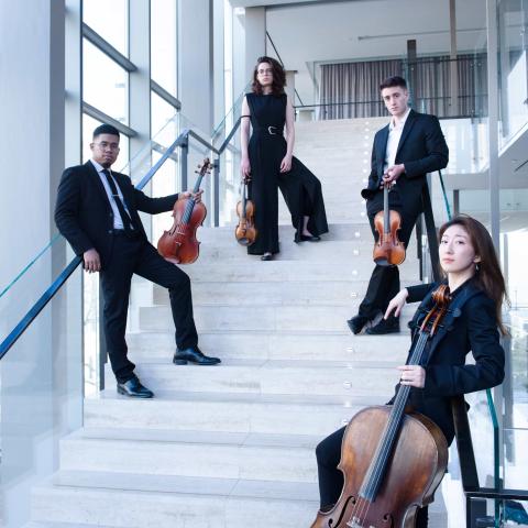 The Dior Quartet stand on white stairs holding their instruments. They lean on the railings and look at the camera. 