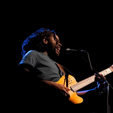Duane holds a guitar. He is performing and the shot is taken from an angle underneath him. He is singing into a micropho