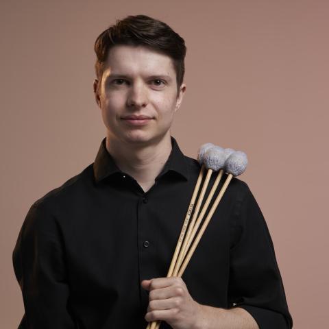 Nolan is pictured here in front of a red background holding mallets. 