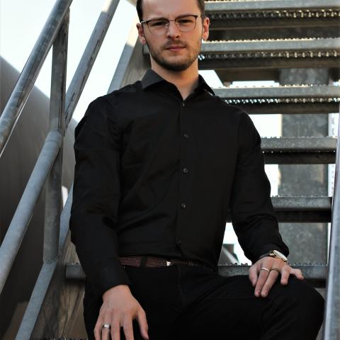 Ethan sits on metal stairs outdoors looking at the camera wearing a dress shirt and dress pants, all black.