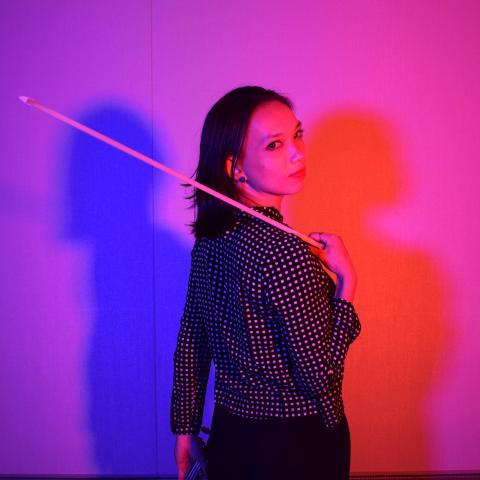 Teagan stands in front of blue and red light with a violin bow.