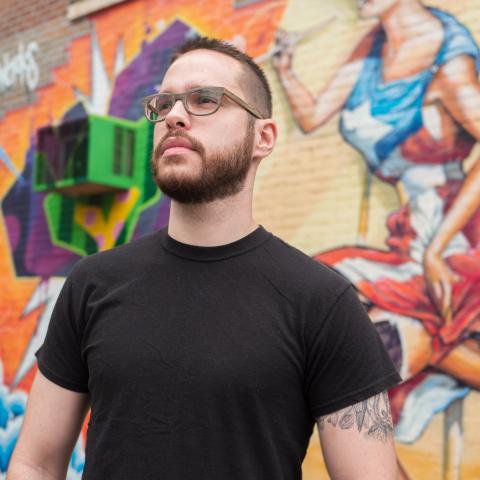 Gabriel stands in a black t-shirt looking to the left in front of a brick wall covered in paintings. 