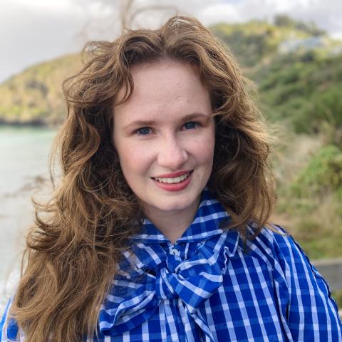Ihlara stands with a blue plaid outfit and hair down in front of an outdoor landscape. She is smiling. 