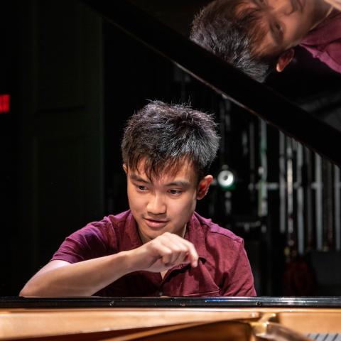 Joey sits at a piano with the lid open. There is a dark background with a red exit sign. We can see his reflection in th